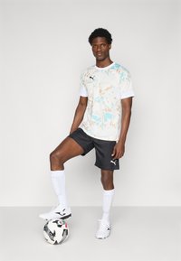 Puma majica v beli, svetlo modri in bež abstraktni vzorec, v kombinaciji z črnimi kratkimi hlačami, belimi nogavicami in belimi nogometnimi kramplji, stoji ob nogometni žogi.