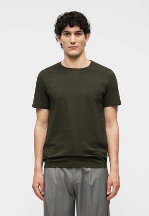 Νέος άνδρας με κοντά σγουρά σκούρα μαλλιά, φορώντας ένα σκουροπράσινο κοντομάνικο μπλουζάκι και ανοιχτόγκρι παντελόνια, στέκεται μπροστά σε απλό φωτεινό φόντο.