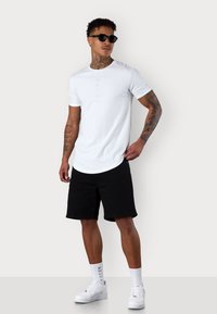 White short-sleeve t-shirt with mint green vertical text, paired with black shorts and white sneakers. Model wearing sunglasses.