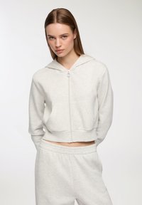 Giovane donna con capelli lisci indossa un giubbino cropped grigio chiaro con zip e pantaloni della tuta abbinati su uno sfondo bianco semplice.