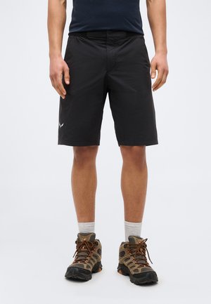 Άνδρας που φοράει μαύρα σορτς μέχρι το γόνατο, ανοιχτό γκρι κάλτσες και καφέ παπούτσια πεζοπορίας, στέκεται μπροστά σε απλό φόντο.