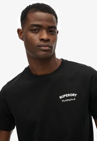 Czarna koszulka z krótkim rękawem wykonana z bawełny, z białym logo "SUPERDRY" i japońskim napisem po lewej stronie klatki piersiowej. Gładka tekstura, regularny krój.