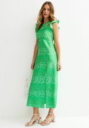 Mujer con vestido midi verde con estampado floral blanco, mangas con volantes, sosteniendo un bolso blanco, y calzando sandalias beige de tacón grueso con tiras.