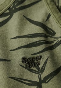 Tkanina w oliwkowym kolorze z ciemnym wzorem liści, z teksturowanym czarnym haftowanym logo "Superdry". Gładka faktura, casualowy design.