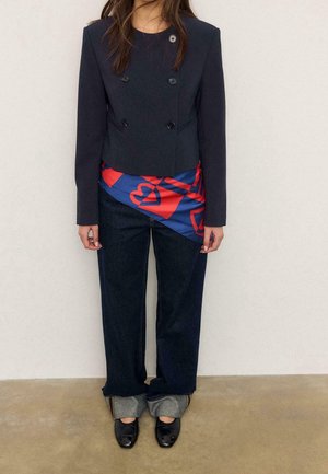 Femme portant un blazer croisé bleu marine, un jean large foncé avec revers, une écharpe rouge et bleue à motifs autour des hanches, et des mocassins noirs.