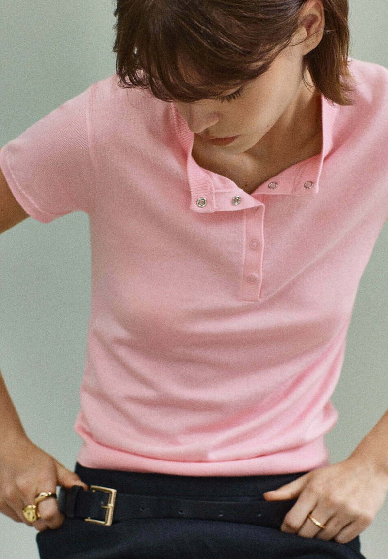 Femme portant un t-shirt rose à manches courtes attachant une ceinture noire autour de sa taille, montrant des bagues à ses doigts.