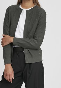 Ciemnozielony kardigan z teksturowanymi kropkami, ściągaczami na mankietach i okrągłym dekoltem, noszony na białej koszuli. Poniżej widoczne czarne spodnie.