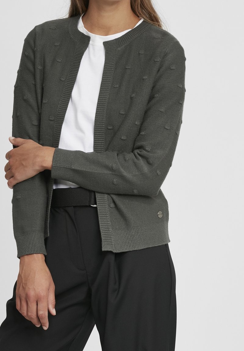 Ciemnozielony kardigan z teksturowanymi kropkami, ściągaczami na mankietach i okrągłym dekoltem, noszony na białej koszuli. Poniżej widoczne czarne spodnie.