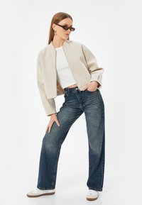 Beige lyhyempi bomber-takki, valkoinen t-paita, leveälahkeiset siniset farkut ja valkoiset lenkkarit, joissa tummat suorakulmaiset aurinkolasit.