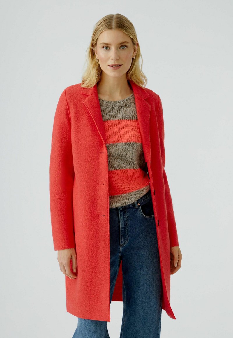 Manteau en laine et alpaga corail avec un design structuré et des revers, porté sur un pull rayé gris et un jean bleu, affichant une coupe droite.