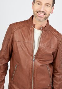 Veste en cuir marron avec des détails matelassés aux épaules, fermetures éclair argentées et poches latérales. Elle présente un col à pression et une texture douce.