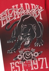 Panthère noire agressive aux dents découvertes au-dessus d'une rose et de fleurs, avec l'inscription "Ed Hardy" et "EST. 1971" sur un tissu rouge.