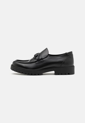 Black leather loafers with a rounded toe, decorative strap, and textured rubber sole featuring a chunky tread pattern.