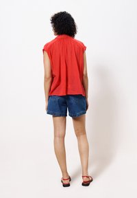 Rote ärmellose Bluse mit gerafftem Rücken, kombiniert mit blauen Denim-Shorts. Das Model trägt schwarze Sandalen mit roten Riemen. Einfaches, entspanntes Design.