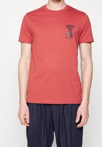 Camiseta de algodón roja con escote redondo y mangas cortas. Presenta una estampado gráfico negro en el pecho. Combinada con pantalones azul marino.