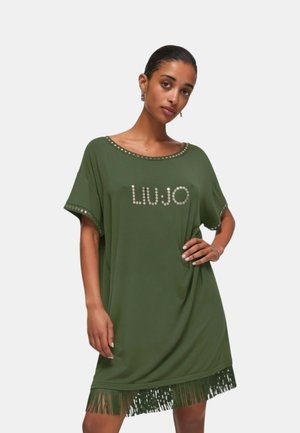 Donna che indossa un abito verde oliva largo con borchie metalliche che formano la scritta "Liu Jo", con rifiniture borchiate e orlo a frange, posando con la mano sui fianchi.