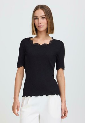 Kvinne med rett, skulderlangt hår iført en svart topp med korte ermer og blonderkant, samt hvite bukser, stående foran en ensfarget bakgrunn.