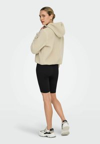 Beige hettejakke i fleece med myk tekstur, kombinert med svarte sykkelshorts. Har en avslappet passform og atletiske joggesko med kontrasterende detaljer.