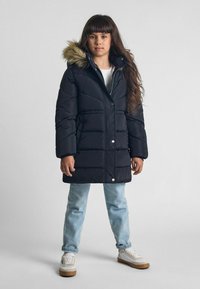 Svart puffjacka med huva i päls, med kviltad textur och silverknappar. Matchad med ljusblå jeans och vita sneakers.