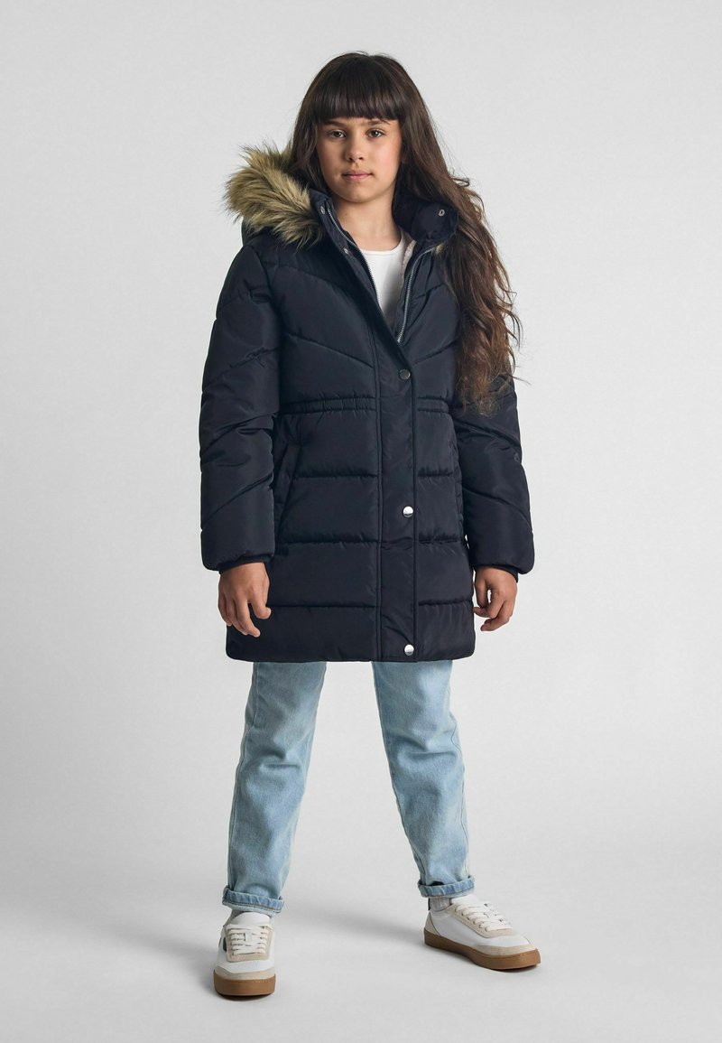 Svart puffjacka med huva i päls, med kviltad textur och silverknappar. Matchad med ljusblå jeans och vita sneakers.