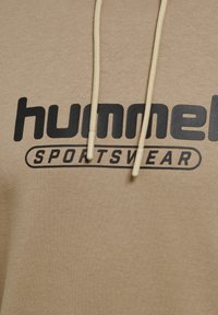 Beżowa bluza z kapturem z napisami "hummel SPORTSWEAR" w grubym czarnym foncie. Posiada sznurki z jasnobeżowymi końcówkami. Miękka, gładka tekstura materiału.