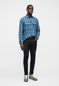 Άνδρας που στέκεται, φοράει μπλε καρό πουκάμισο με κουμπιά, σκούρο τζιν, καφέ ζώνη και πράσινα-λευκά αθλητικά παπούτσια, με το χέρι στην τσέπη, μπροστά σε λευκό φόντο.