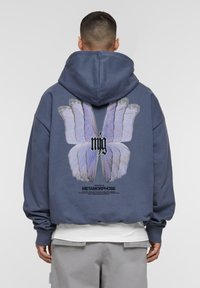 Blaues oversized Hoodie mit einem großen Schmetterlings-Graphic in Lila auf der Rückseite, mit Text darunter. Aus Baumwollmaterial, weiche Textur, gerippte Bündchen.