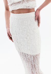 Jupe en dentelle blanche présentant des motifs floraux complexes, une coupe ajustée et une silhouette longueur genou avec une section inférieure transparente.
