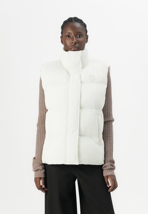 White puffer vest with high collar, featuring horizontal quilted design and front pockets, layered over a ribbed brown top.