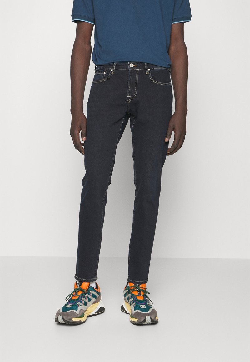 Homme portant un jean slim foncé, un polo bleu et des baskets athlétiques multicolores, debout devant un fond blanc uni.