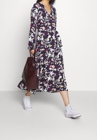 Dlouhá, splývavá šaty s tmavě fialovým základem a květinovým vzorem v růžové, bílé a oranžové barvě. Kombinované s bílými teniskami a hnědou kabelkou.