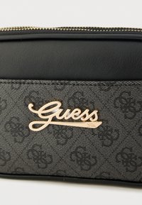 Čierna syntetická peňaženka s textúrovaným sivým vzorom a zlatým logom "Guess" na prednej strane. Zipsové zapínanie na vrchu.