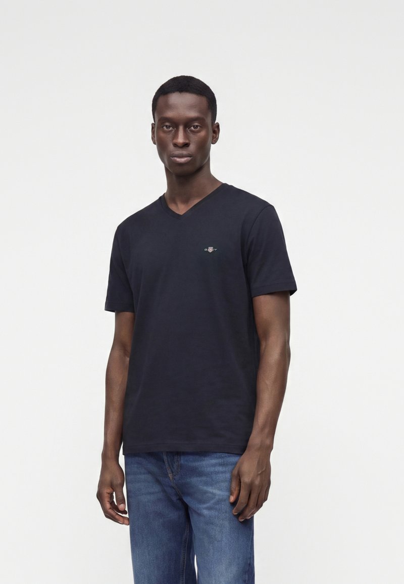 Homme aux cheveux courts portant un t-shirt noir à col en V et un jean bleu, debout devant un fond blanc uni.