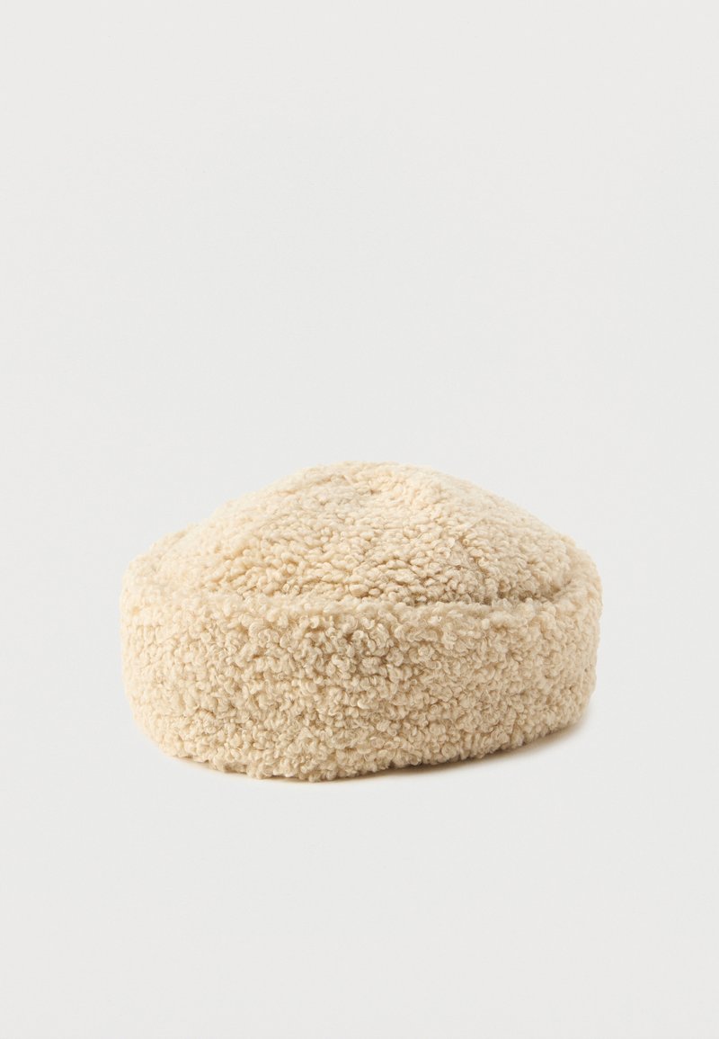 Beige runder Hut aus strukturiertem, flauschigem Material. Er hat eine glatte, leicht erhöhte Oberseite mit einer dezenten Naht um den Boden.