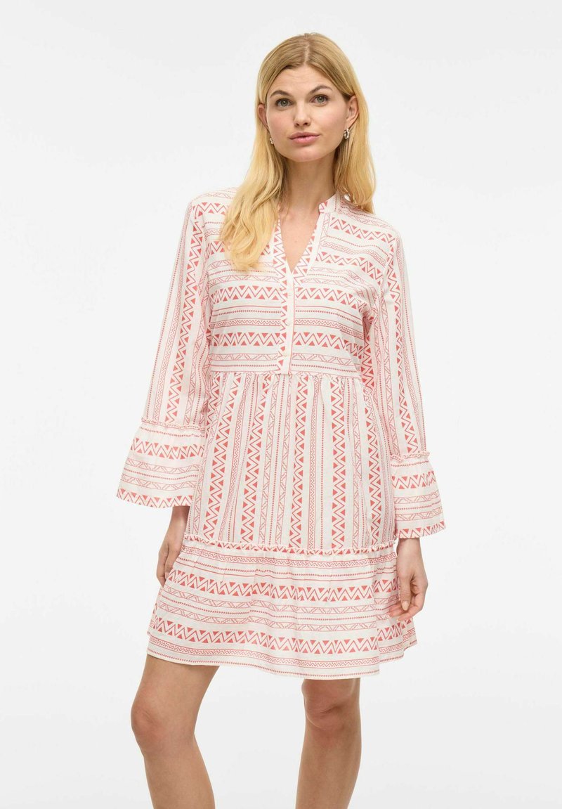 Robe blanche avec des motifs géométriques rouges, manches évasées, col en V et taille froncée. Le tissu semble léger avec une texture douce.