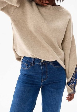 Person trägt beigefarbenen Strickpullover mit gemusterten Bündchen und hoch taillierte blaue Jeans, posiert mit Hand nahe dem Gesicht.