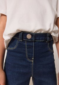 Tamno plave traperice s zlatim šavovima, metalnim zatvaračem i džepovima. Uparene s jednostavnom bijelom majicom.