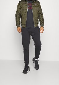 Man in een olijfgroen pufferjack, zwart graphic T-shirt, zwarte joggers, witte sokken en zwarte Nike sneakers loopt over een grijze vloer.