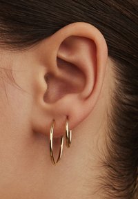 Des boucles d'oreilles en or présentées en gros plan sur une oreille. Les boucles d'oreilles ont une finition lisse et brillante ainsi qu'un design circulaire.