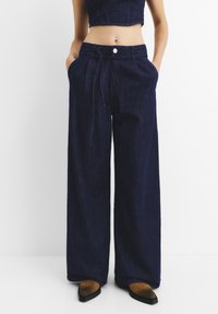 Široke temnoplave jeans hlače z elastičnim pasom in sprednjimi žepi, ki se ponašajo z gladko teksturo in klasičnim gumbom za zapiranje.