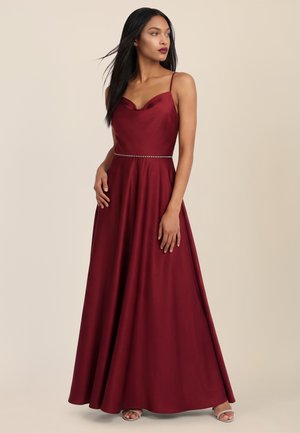 Frau, die ein ärmelloses bordeauxrotes Abendkleid mit dünnen Trägern und einem perlenbesetzten Taillenband trägt, steht vor einem einfarbigen beigen Hintergrund.