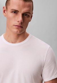 Jonge man met kort haar die een effen wit T-shirt met ronde hals draagt tegen een effen grijze achtergrond.