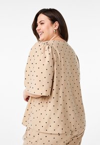Beige Oberteil mit Polka-Dots und kurzen Puffärmeln, mit schwarzen Punkten und weicher Textur, gestaltet mit einer entspannten Passform und sichtbaren Nähdetails.