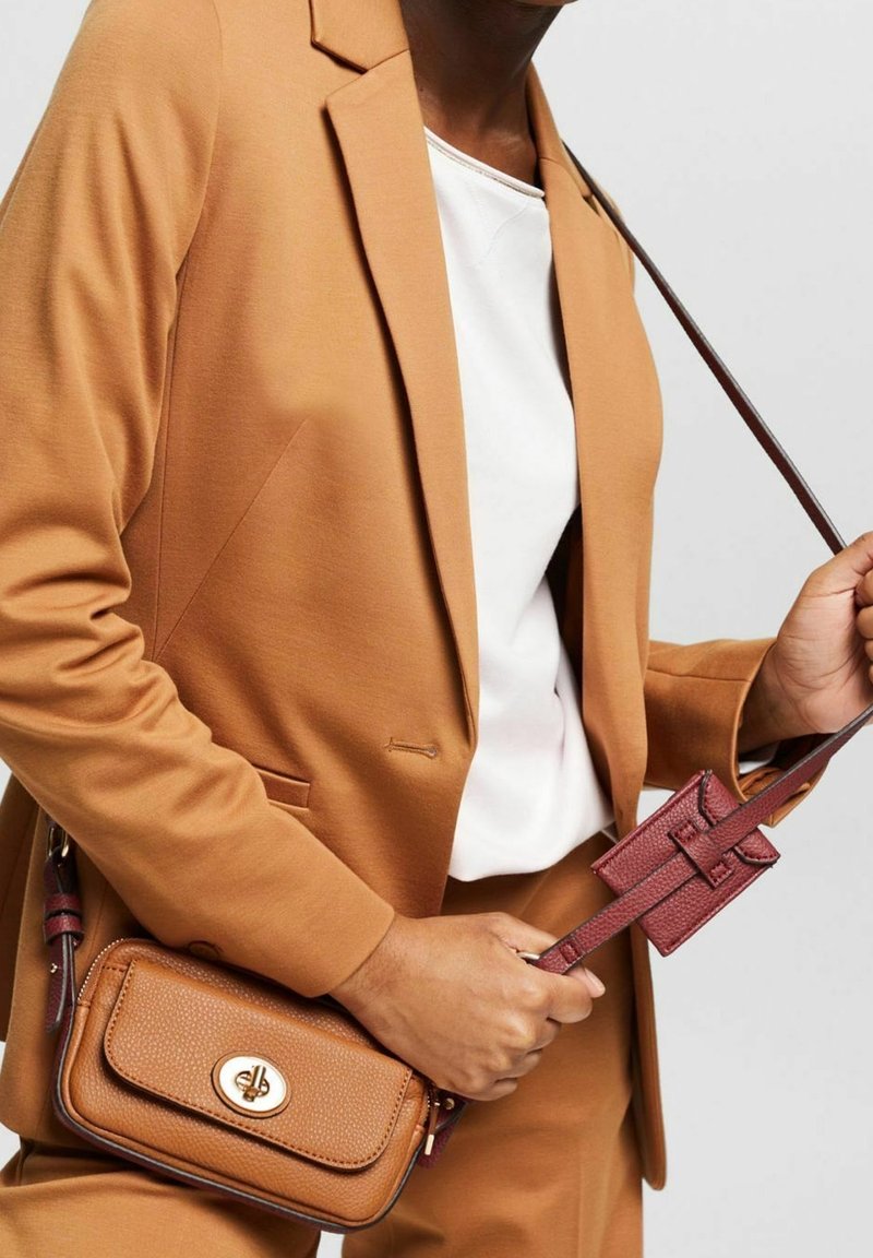 Blazer e pantaloni beige; modella che tiene una borsa a tracolla in pelle marrone con dettagli dorati, decorata con un piccolo accessorio rosso.