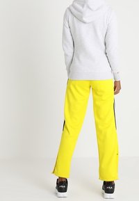 Felpa grigia con cappuccio, abbinata a pantaloni da tuta gialli brillanti con strisce nere sui lati e una piccola fessura alle caviglie. Sneakers nere.