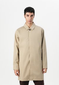 Casaco bege, longo, com colarinho, fechamento com um único botão e bolsos laterais, confeccionado em um tecido liso e texturizado.