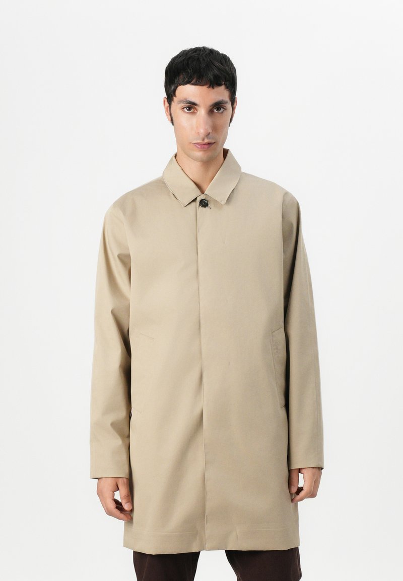 Casaco bege, longo, com colarinho, fechamento com um único botão e bolsos laterais, confeccionado em um tecido liso e texturizado.