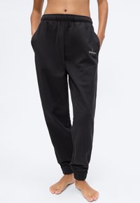 Άτομο που φοράει μαύρο παντελόνι φόρμας Calvin Klein με ελαστικές μανσέτες και χέρια στις τσέπες, ξυπόλητο, μπροστά σε απλό φόντο.