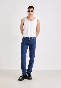 Biely bezrukávový top, modré džínsy a čierne šnurovacie topánky. Modelka so slnečnými okuliarmi stojí na neutrálnom pozadí.