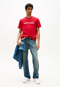 Rotes Baumwoll-T-Shirt mit weißem "TOMMY JEANS"-Logo, kombiniert mit blauen Jeans. Das Modell hält eine blaue Jeansjacke und trägt weiße Sneakers.
